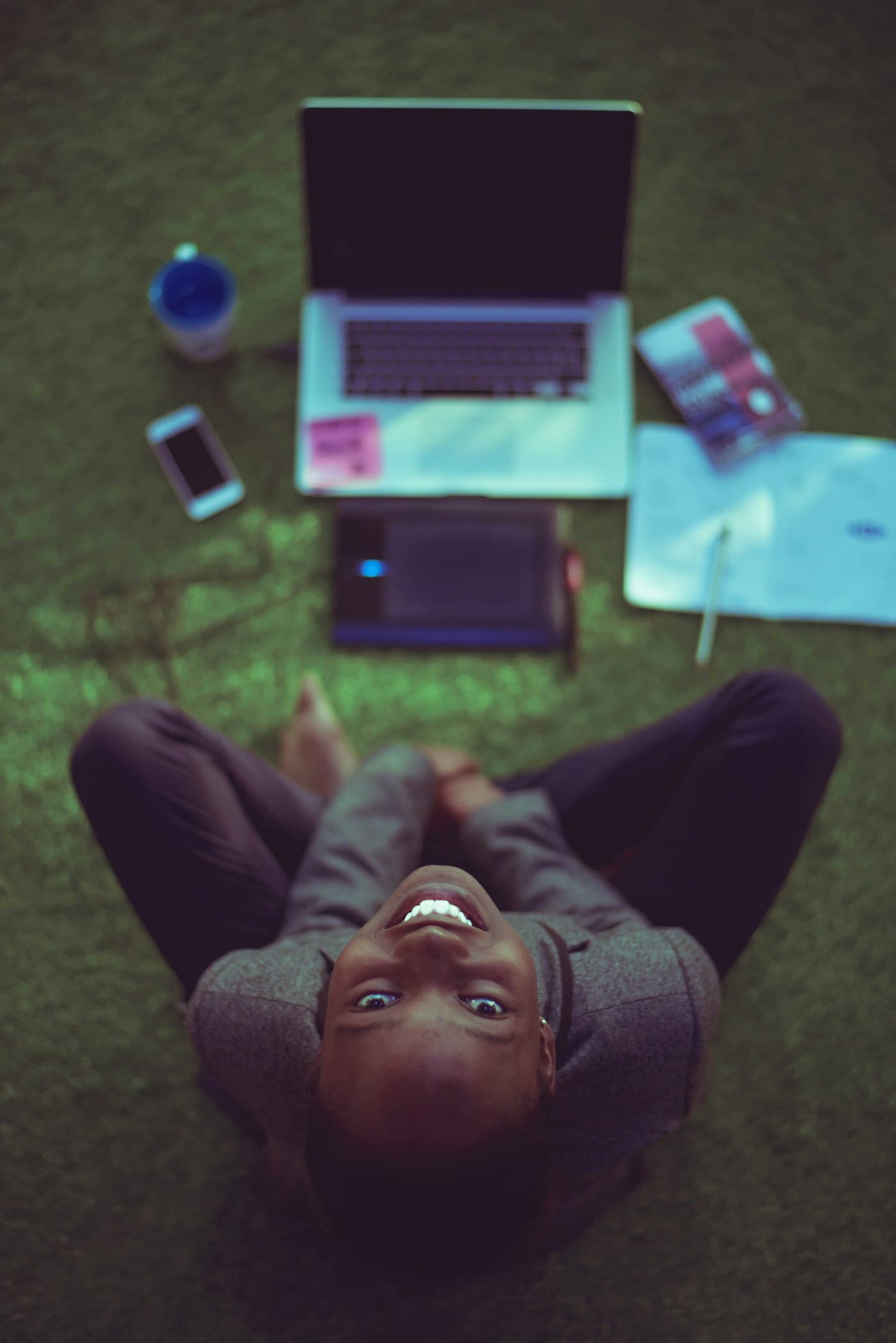 A smiling woman lying on the grass beside her laptop and phone